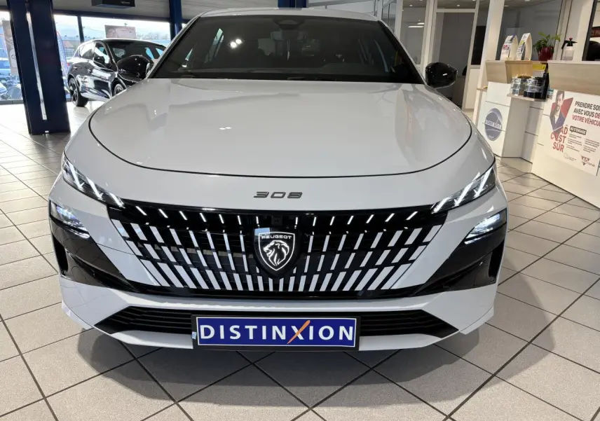 Vue de face d'une Peugeot 308 blanche avec calandre noire à motifs verticaux et logo lion lumineux, en showroom.