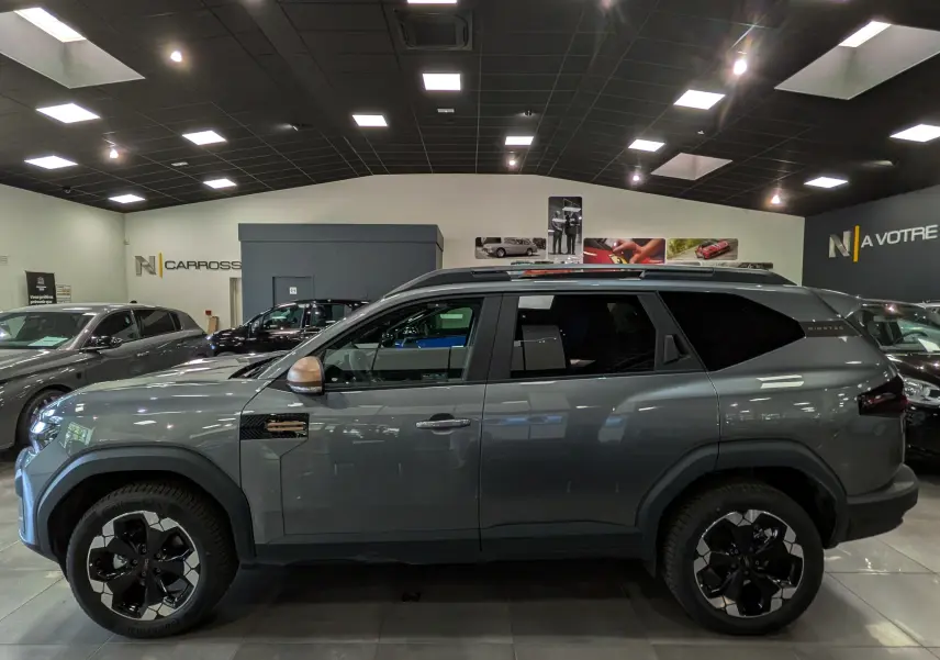 Vue de profil côté gauche du Dacia Bigster Hybrid-G 150 4x4 gris schiste avec jantes bicolores et toit noir en showroom.