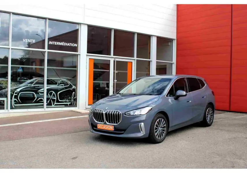 BMW Série 2 Active Tourer gris vue 3/4 avant avec calandre chromée, stationnée devant un bâtiment orange et blanc.