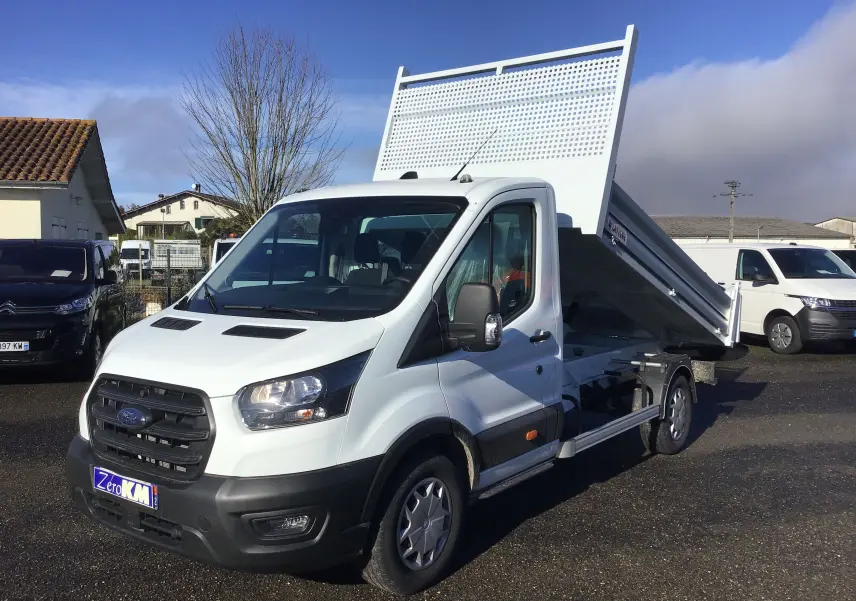 Vue 3/4 avant droite d'un Ford Transit blanc avec benne relevée et cabine simple sur un parking extérieur.