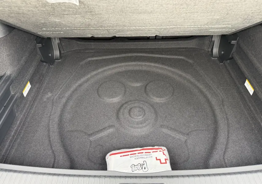 Coffre ouvert vue de dessus, tapis de sol gris avec espace pour roue de secours, petit kit de secours blanc visible.