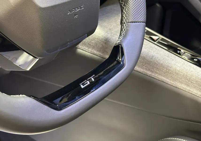 Gros plan sur le volant cuir noir avec logo GT du Peugeot 3008 III Hybrid, intérieur gris clair et détails modernes.