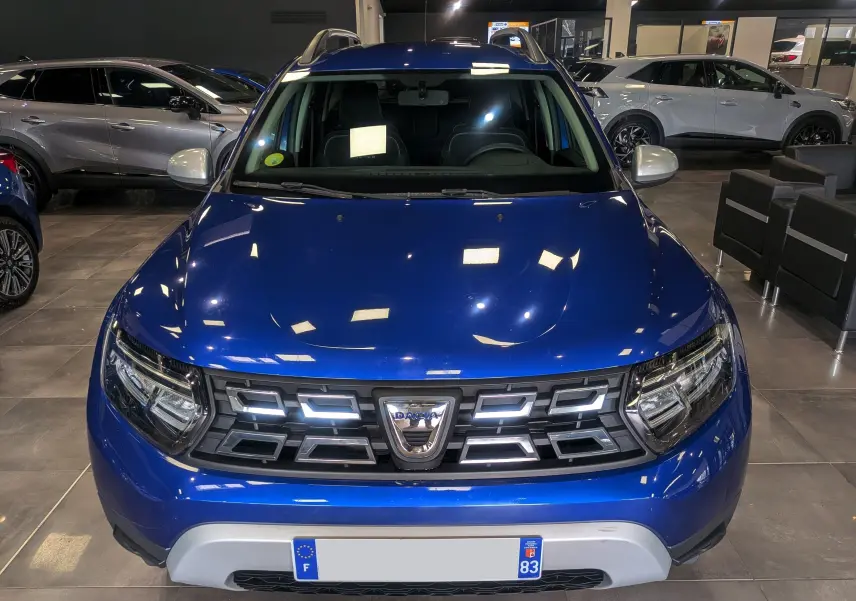 Vue avant d'un Dacia Duster 2022 Bleu Iron en showroom, avec calandre distinctive et phares LED allumés.