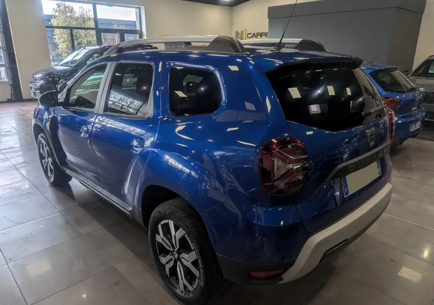 Vue 3/4 arrière droite du Dacia Duster 2022 Bleu Iron avec jantes alliage bicolores et barres de toit noires.