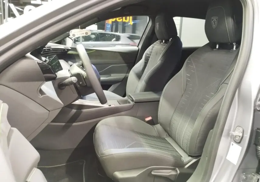 Vue intérieure côté conducteur de la Peugeot 308 gris Artense, sièges noirs avec surpiqûres vert Adamite et logo Peugeot brodé.