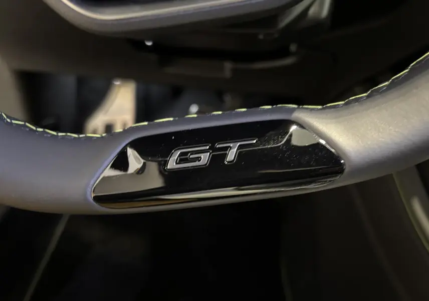 Gros plan sur le volant noir avec surpiqûres vert Adamite et logo GT brillant de la Peugeot 308 Hybrid 2026.