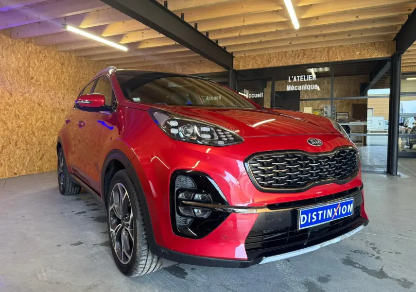 KIA Sportage rouge 2019 en 3/4 avant droit, avec calandre noire et jantes alliage visibles en intérieur d'atelier.