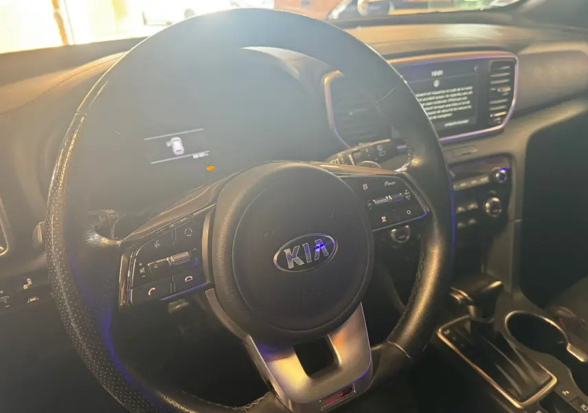 Vue intérieure centrée sur le volant noir perforé du Kia Sportage 1.6 CRDI, avec tableau de bord et écran tactile en arrière-plan.
