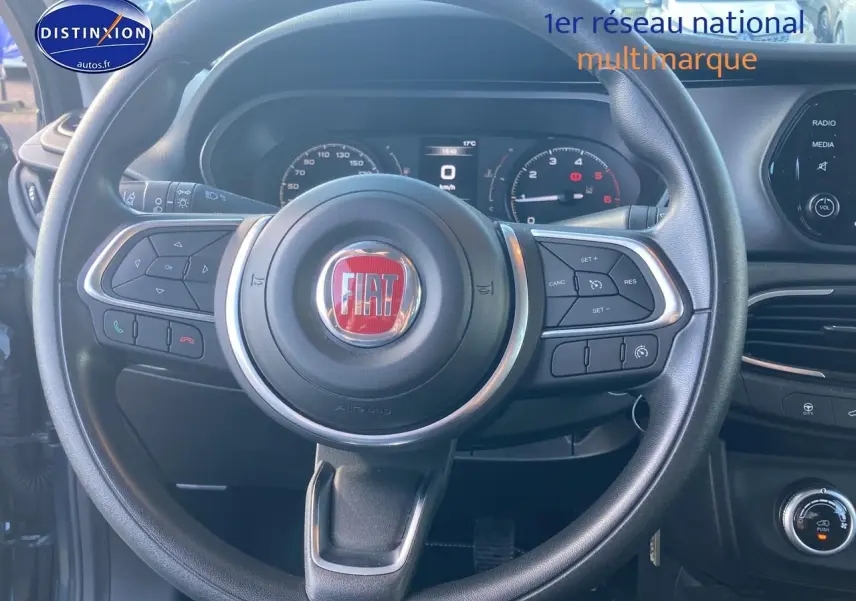 Vue rapprochée du volant multifonction noir avec logo FIAT rouge et tableau de bord numérique d'une Fiat Tipo 2024.