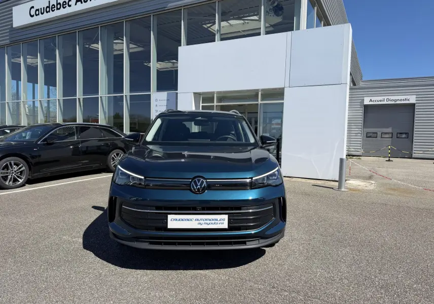 Vue avant d'un Volkswagen Tiguan bleu métallisé garé devant un concessionnaire avec calandre noire et logo VW central.