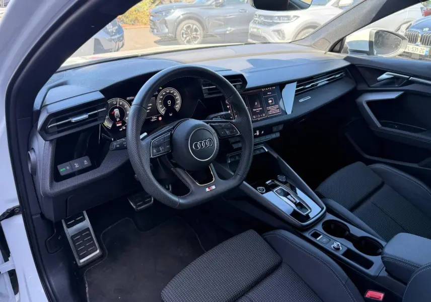 Intérieur Audi A3 Sportback blanc, vue côté conducteur montrant tableau de bord noir et volant sport multifonction.