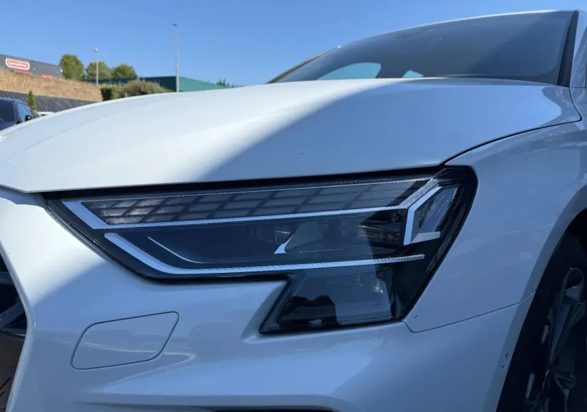 Gros plan sur le phare avant droit d'une Audi A3 Sportback blanc Glacier métallisé, avec jante alu visible en arrière-plan.