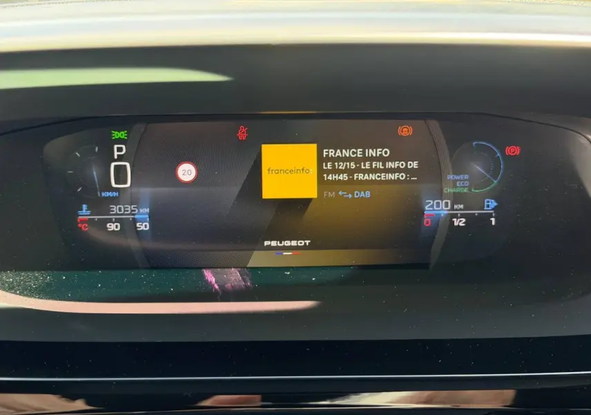 Affichage numérique du tableau de bord Peugeot 408 Hybrid 145ch GT, montrant la radio France Info et les indicateurs de vitesse et batterie.