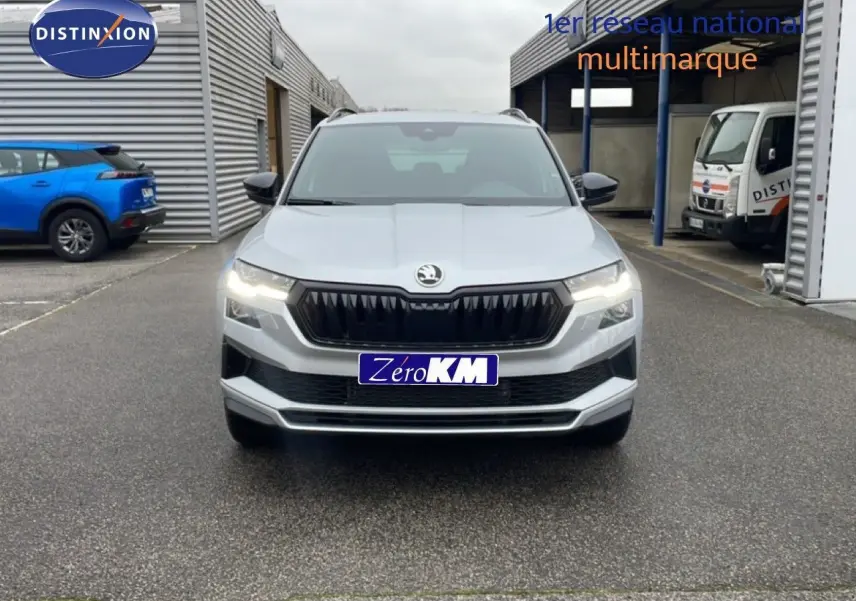 Vue frontale d'un Skoda Karoq gris argent métal 2025 avec calandre noire et feux allumés sur parking.