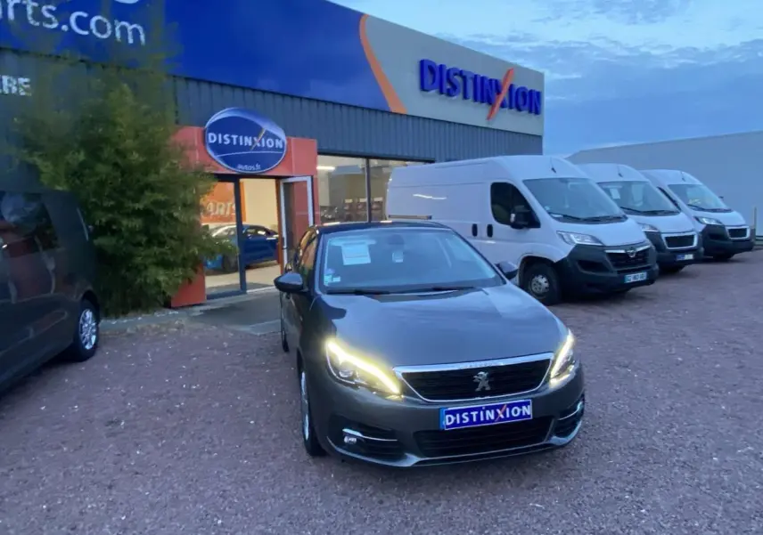 Peugeot 308 gris clair vue 3/4 avant avec phares allumés, stationnée devant un concessionnaire Distinxion.