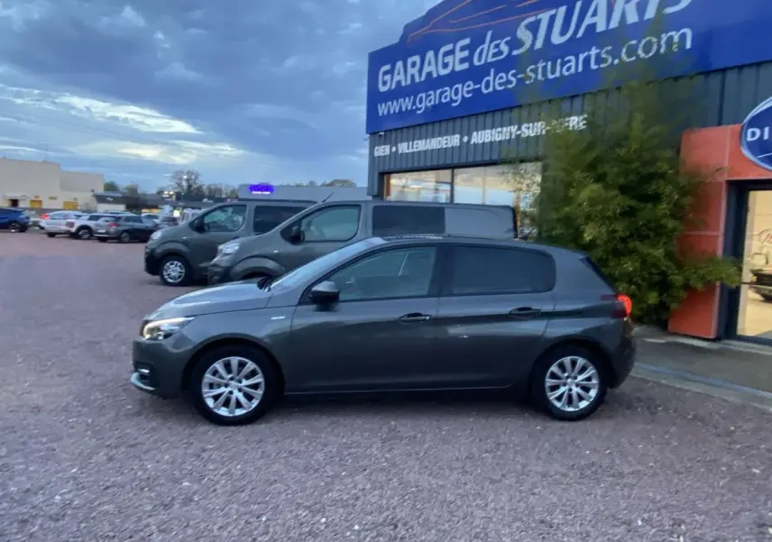 Peugeot 308 gris clair vue de profil côté gauche, avec jantes aluminium 16 pouces, stationnée devant un garage.