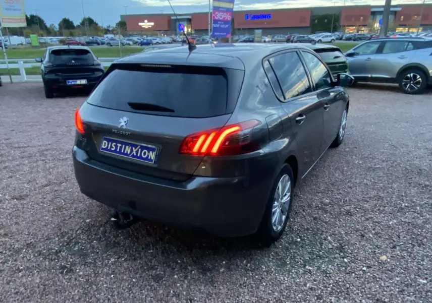 Peugeot 308 gris clair vue 3/4 arrière droit, feux arrière allumés, vitres surteintées, jantes aluminium 16 pouces.