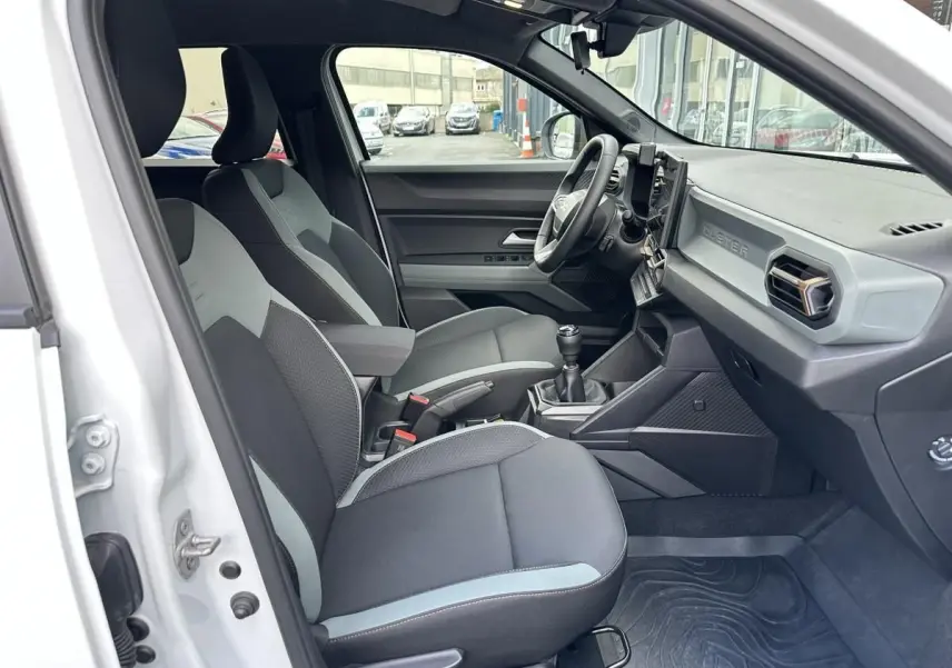 Vue intérieure côté conducteur du Dacia Duster blanc glacier 2025, montrant sièges tissu bicolore et tableau de bord moderne.