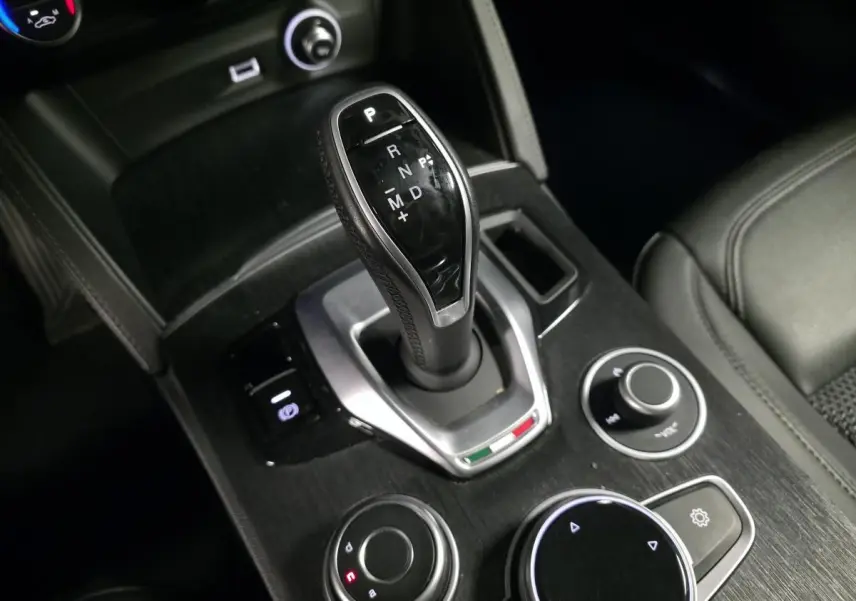 Gros plan sur la console centrale noire de l'Alfa Romeo Stelvio 2.2 D avec levier de vitesse automatique et commandes rotatives.