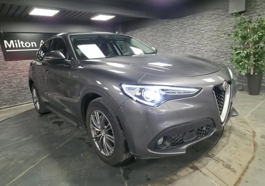 Vue 3/4 avant gauche d'un Alfa Romeo Stelvio gris métallisé avec jantes alliage 18 pouces et phares LED allumés.