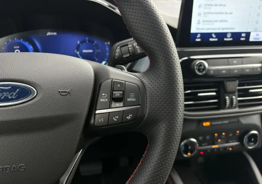 Gros plan sur le volant perforé noir avec surpiqûres rouges du Ford Kuga 2023, tableau de bord numérique et écran tactile visible.