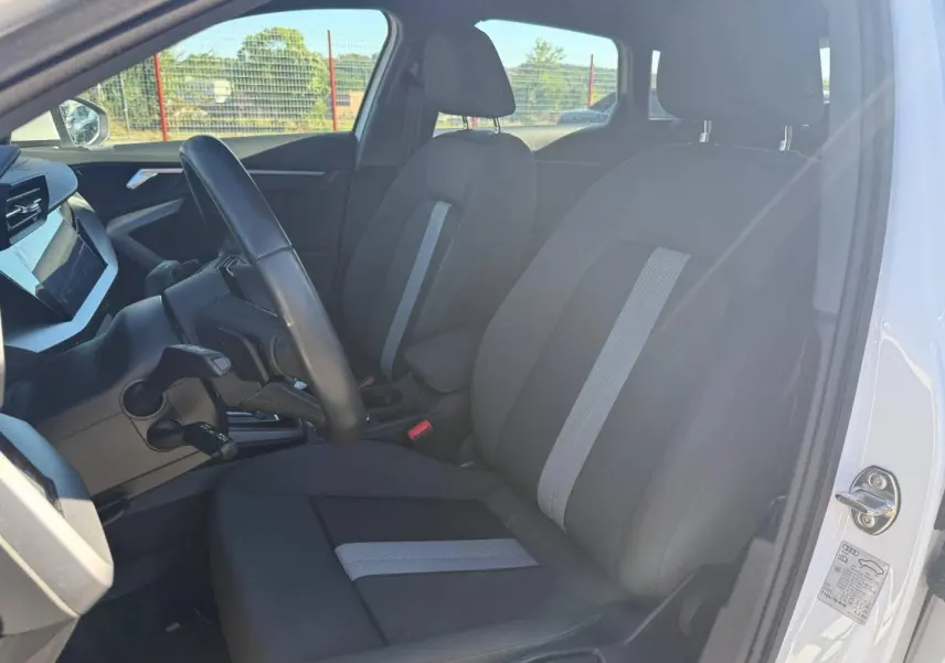 Vue intérieure côté conducteur de l'Audi A3 Sportback blanc Arkona, montrant les sièges tissu noir avec bande centrale grise.