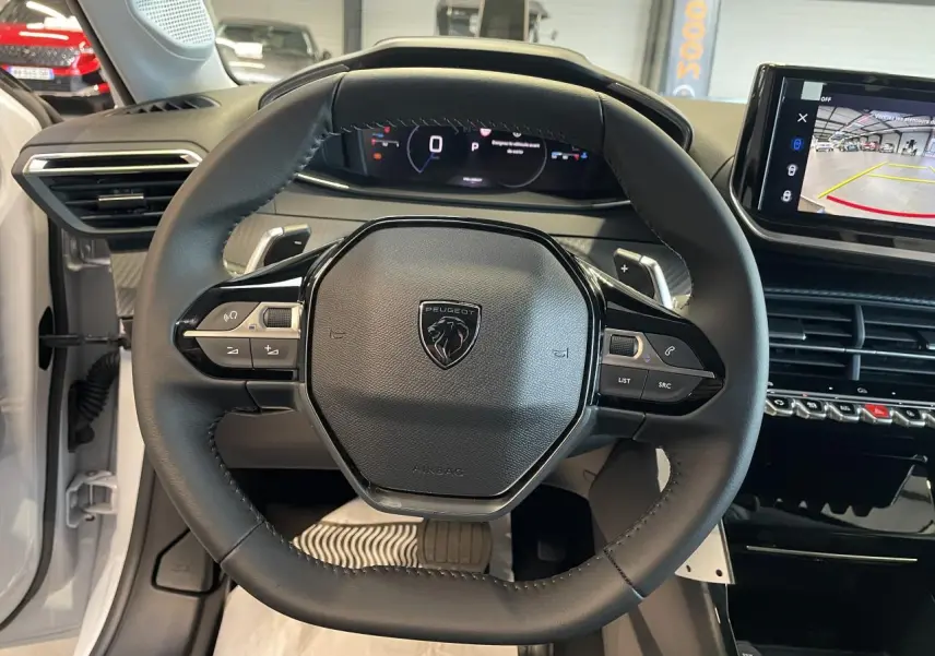 Vue intérieure centrée sur le volant cuir noir avec logo Peugeot, tableau de bord digital et écran tactile en arrière-plan.