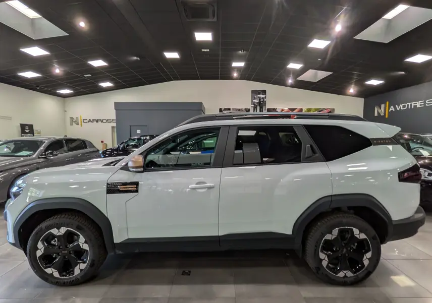 Vue de profil côté gauche du Dacia Bigster Hybrid-G 150 4x4 blanc Glacier avec jantes bi-ton et toit noir en showroom.