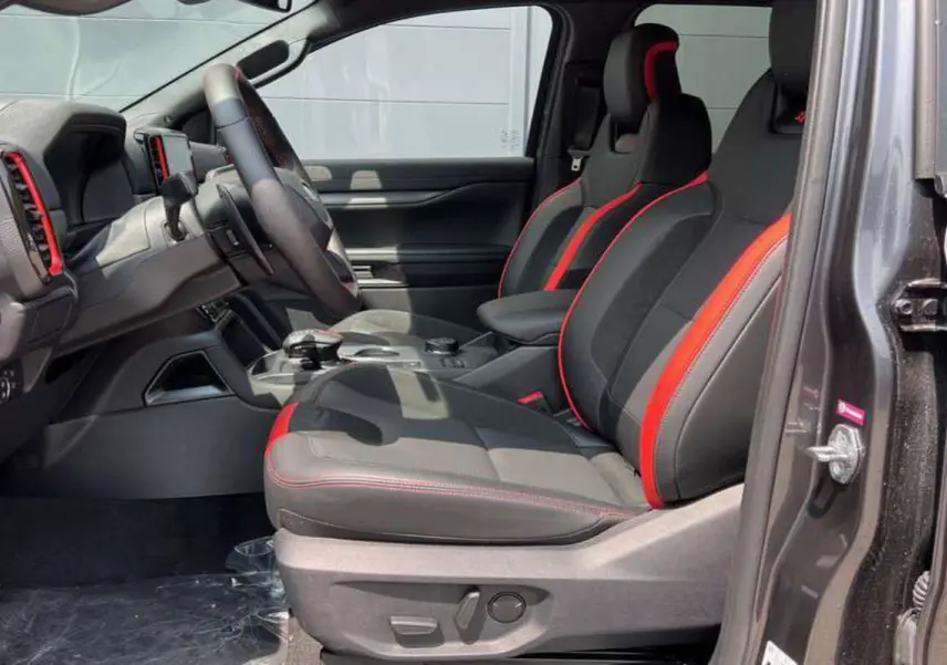 Vue intérieure côté gauche du Ford Ranger Raptor 2025, sièges sport noirs avec surpiqûres rouges et volant cuir multifonctions.