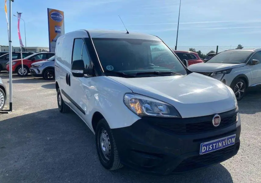 Fiat Doblo blanc vue 3/4 avant droit avec pare-chocs noir non peint et porte latérale droite visible.