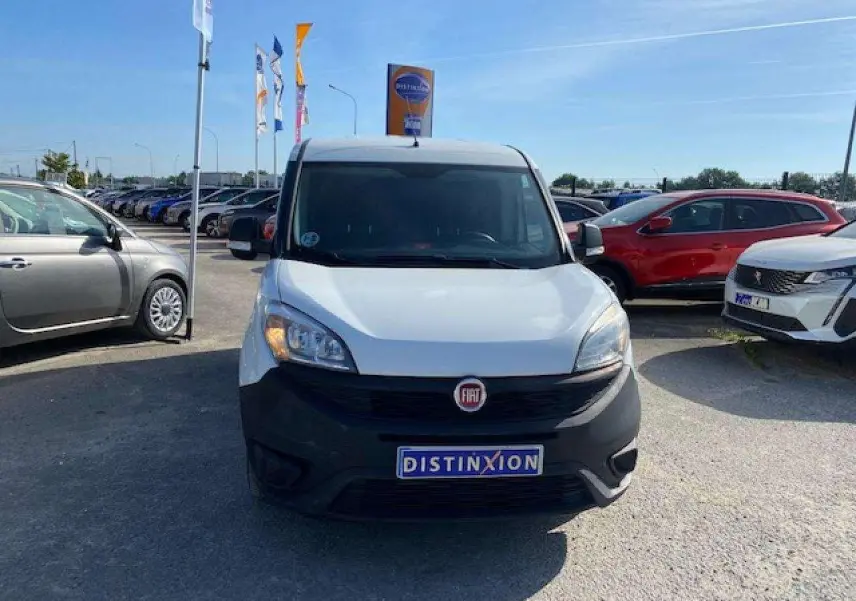 Vue avant d'un FIAT Doblo blanc avec pare-chocs noir mat, stationné sur un parking en plein jour.