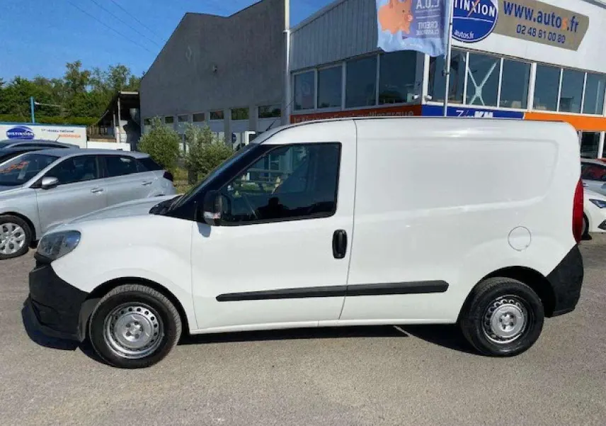 Vue de profil côté gauche d’un FIAT Doblo blanc fourgon tôlé avec pare-chocs noirs et porte latérale droite visible en arrière.