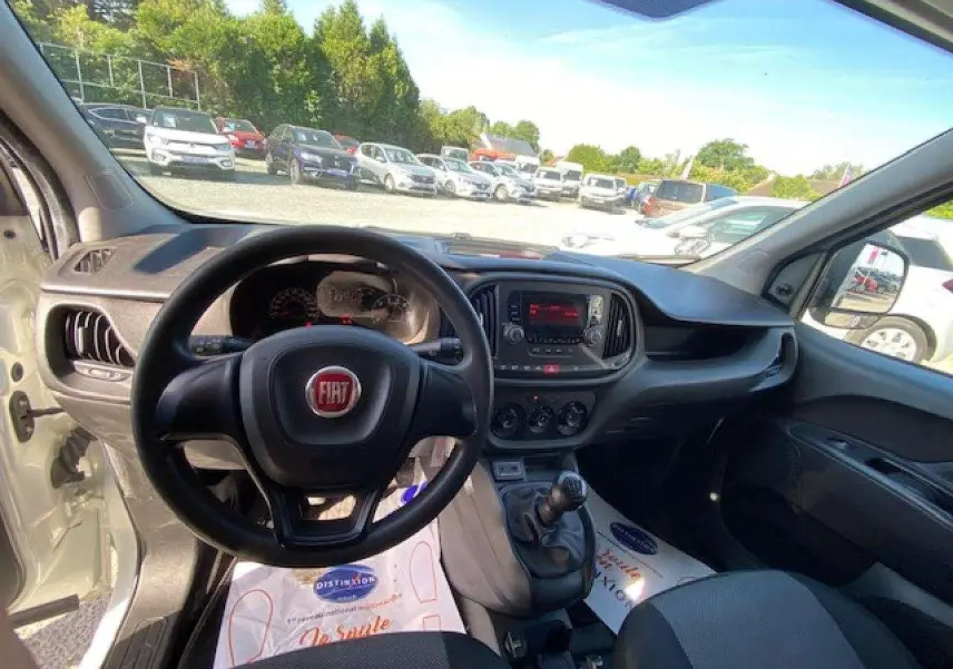Vue intérieure du poste de conduite du Fiat Doblo blanc de 2017, avec volant, tableau de bord et levier de vitesse manuelle visibles.