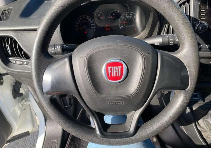 Vue rapprochée du volant noir avec logo rouge FIAT du fourgon blanc Doblo, tableau de bord et levier de vitesses visibles.