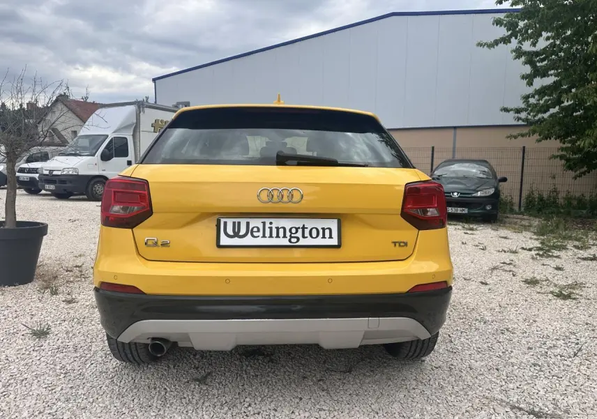 Vue arrière d'un Audi Q2 jaune avec feux rouges et sortie d'échappement chromée visible sur un sol gravillonné.
