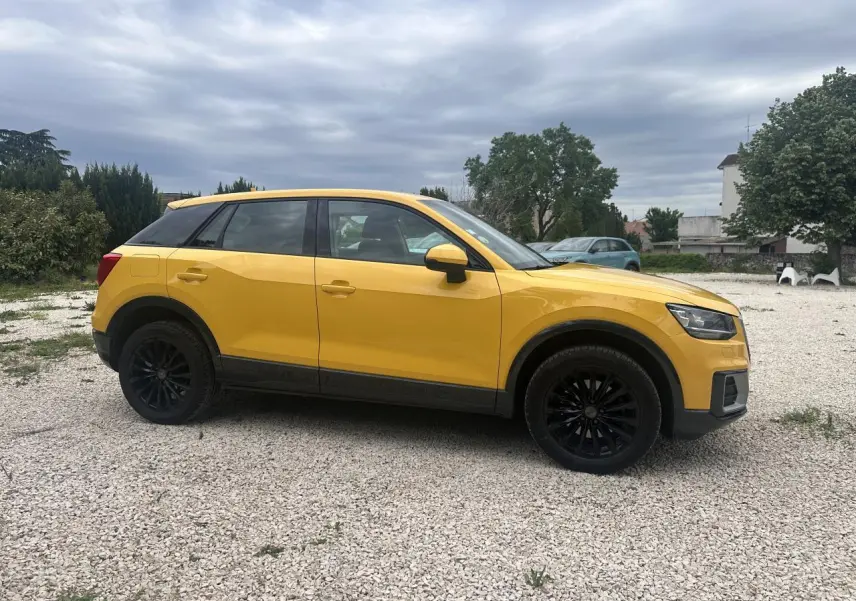 Audi Q2 jaune vue de profil côté gauche avec jantes noires et toit noir sur sol gravillonné.