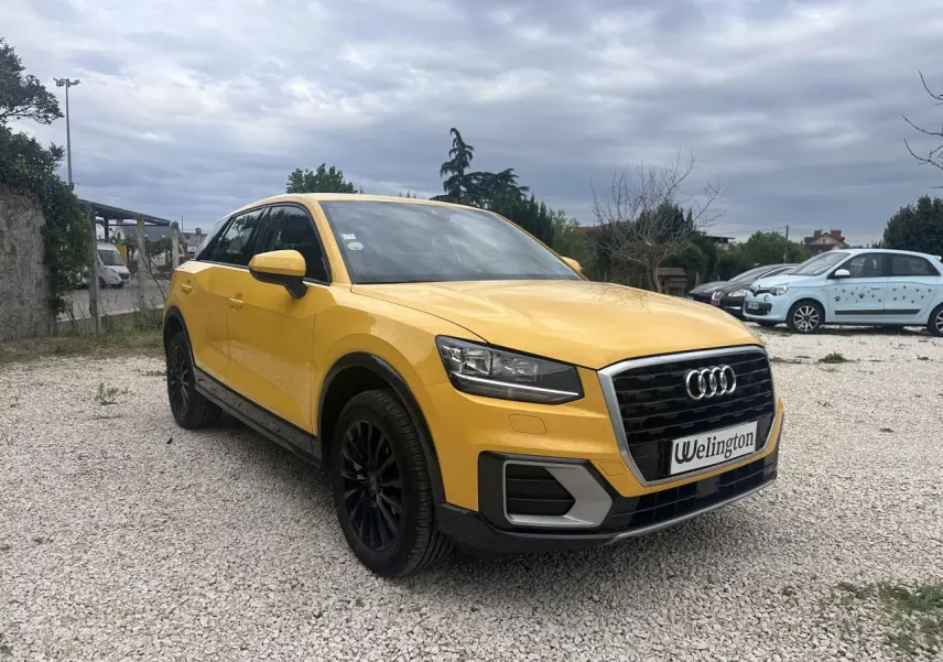 Audi Q2 jaune vue 3/4 avant droit avec calandre noire et jantes noires sur parking gravillonné.