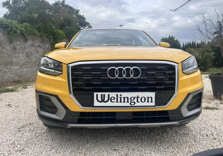 Vue frontale d'un Audi Q2 jaune avec calandre noire et contours chromés, sur sol gravillonné.