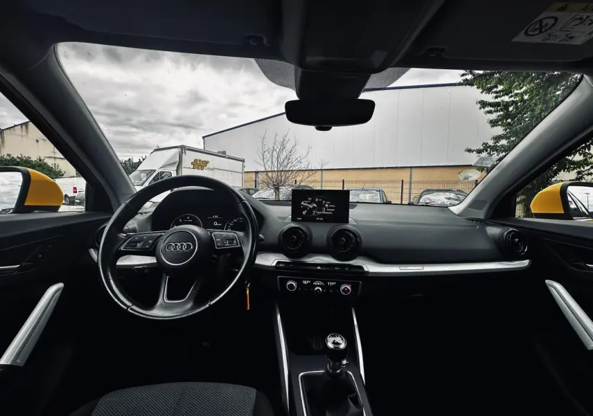 Vue intérieure avant de l'Audi Q2 jaune, volant multifonction, tableau de bord moderne avec écran central et boîte manuelle.