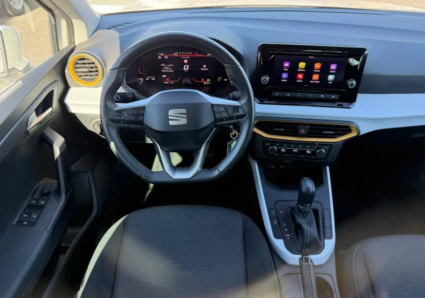 Vue intérieure avant du tableau de bord de la SEAT Arona 2025 avec volant cuir, écran tactile et accents jaunes autour des aérateurs.
