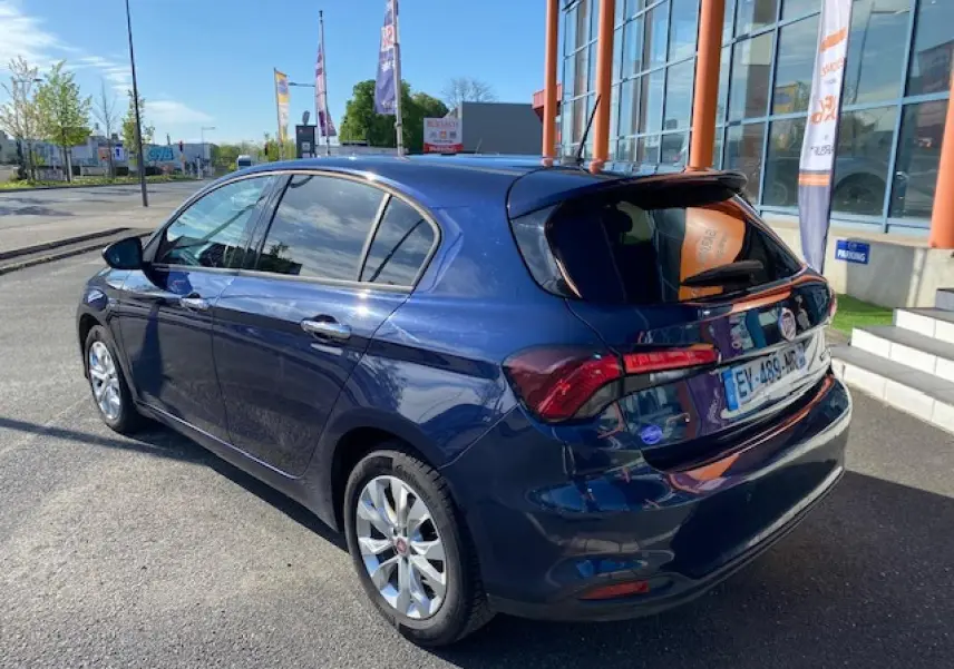 Vue 3/4 arrière droite d'une Fiat Tipo bleu métallisé garée devant un bâtiment avec vitres surteintées et jantes argentées.