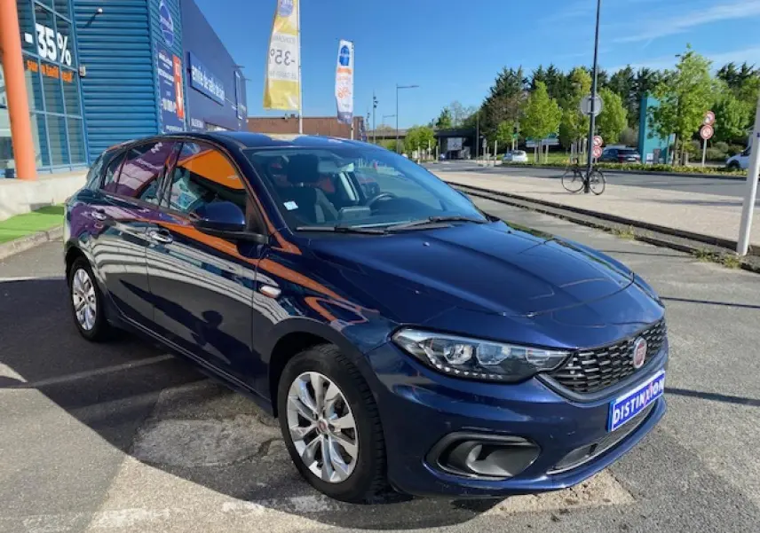 Vue 3/4 avant droite d'une FIAT Tipo bleu métallisé garée devant un bâtiment commercial par temps ensoleillé.