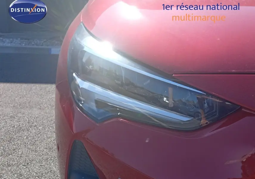 Gros plan sur le phare avant gauche d’une Opel Corsa rouge métal avec toit noir, sous un fort éclairage solaire.