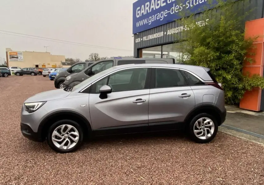 Opel Crossland X gris métallisé en profil droit, avec jantes alliage 16'' et vitres surteintées, stationné devant un garage.