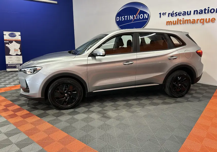 Vue de profil côté gauche du MG ZS 1.5 L Hybrid+ 2025 en Cosmic Silver avec jantes noires, dans un showroom.