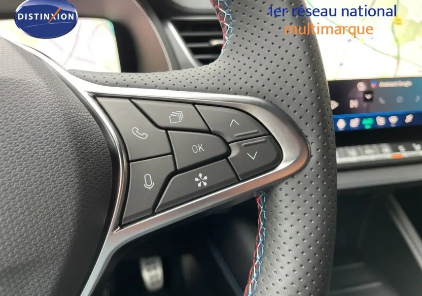 Gros plan sur les commandes au volant en cuir perforé gris avec surpiqûres tricolores, tableau de bord numérique en arrière-plan.