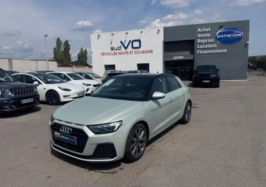 Audi A1 Sportback gris vue 3/4 avant droit, avec toit noir, stationnée devant un garage de véhicules neufs et occasions.
