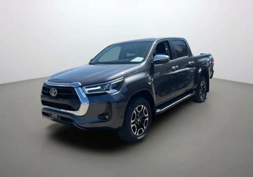 Toyota Hilux 4x4 gris atlas en 3/4 avant droit, avec jantes alu 18 pouces et marchepieds chromés visibles.