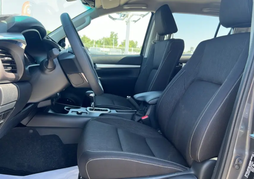 Vue intérieure côté conducteur du Toyota Hilux 2026 gris atlas, sièges tissu noirs avec coutures blanches et console centrale.