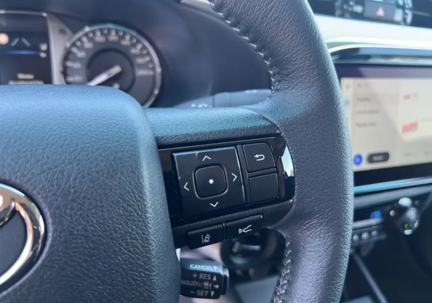 Gros plan sur le volant cuir multifonctions noir du Toyota Hilux 2026 avec tableau de bord et écran tactile visible en arrière-plan.